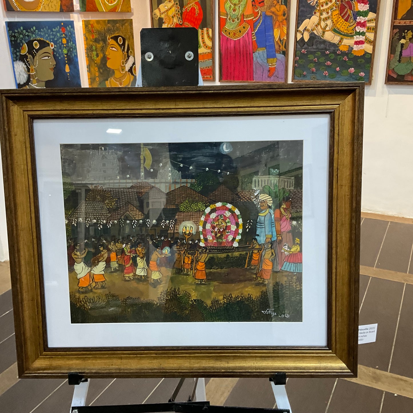 Framed artwork of a colorful scene with people and an elephant, displayed on a stand.